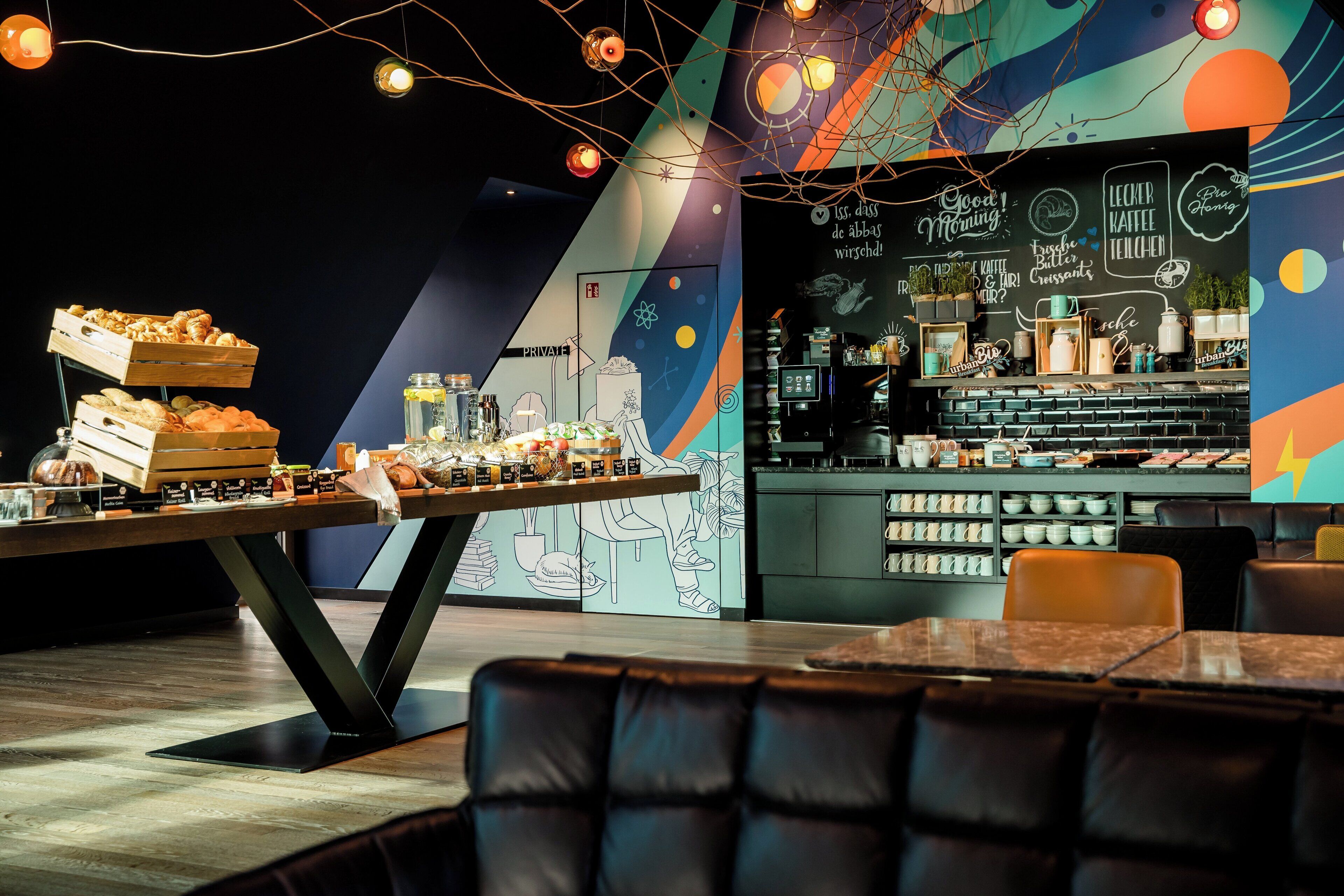 Una cafeteria amb una barra plena de pastissos i cafè, un mural amb obra d'art inspirada en l'espai, i una zona de seients amb butaques de cuir.