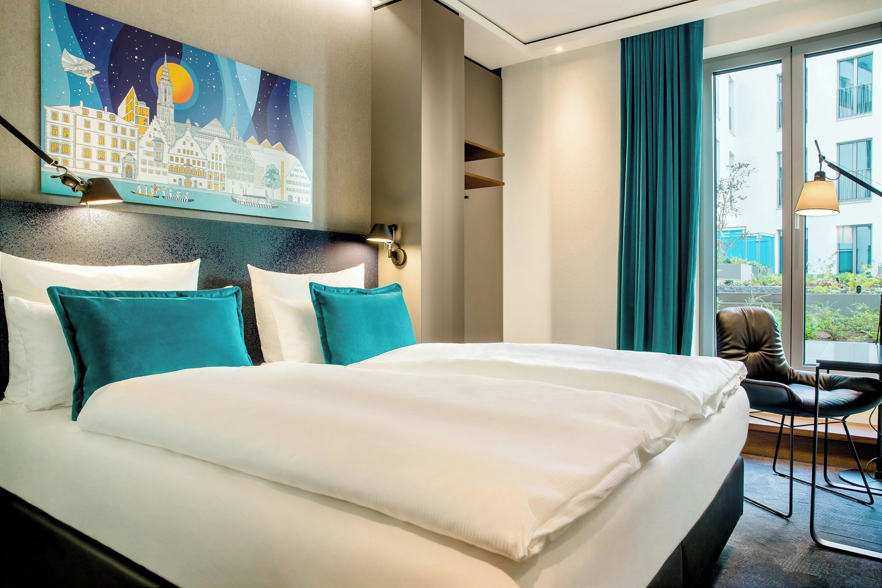 A hotel room with a large bed, teal pillows, a cityscape painting, a view of the outdoors, and a chair by the window.