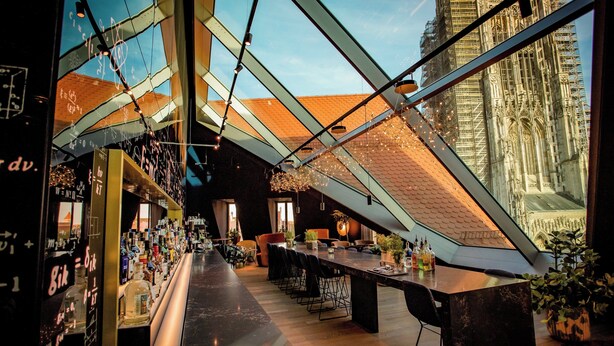 A modern bar area with a high ceiling, exposed beams, and a view of a historic building through large windows.