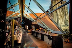 Zona bar moderna con soffitto alto, travi a vista e vista su un edificio storico attraverso ampie finestre.