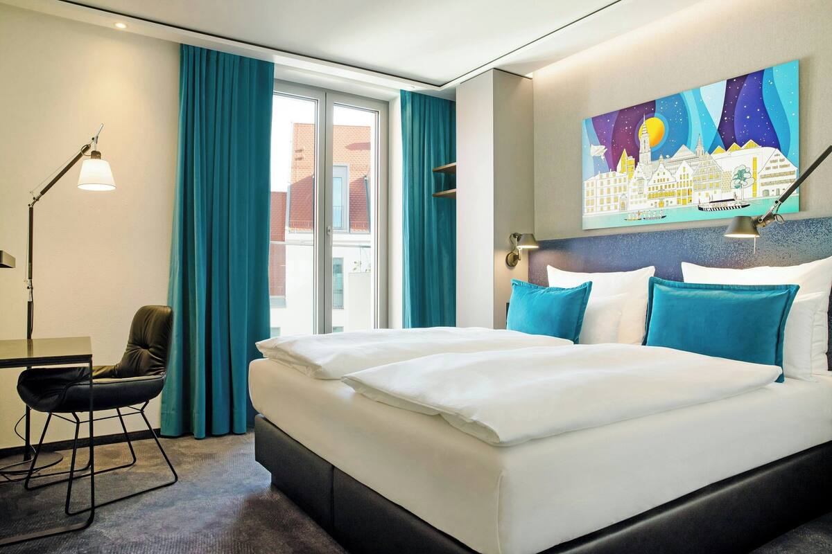 A hotel room with a large bed, a desk, a chair, a desk lamp, and a cityscape painting on the wall.