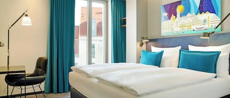A hotel room with a large bed, a desk, a chair, a desk lamp, and a cityscape painting on the wall.