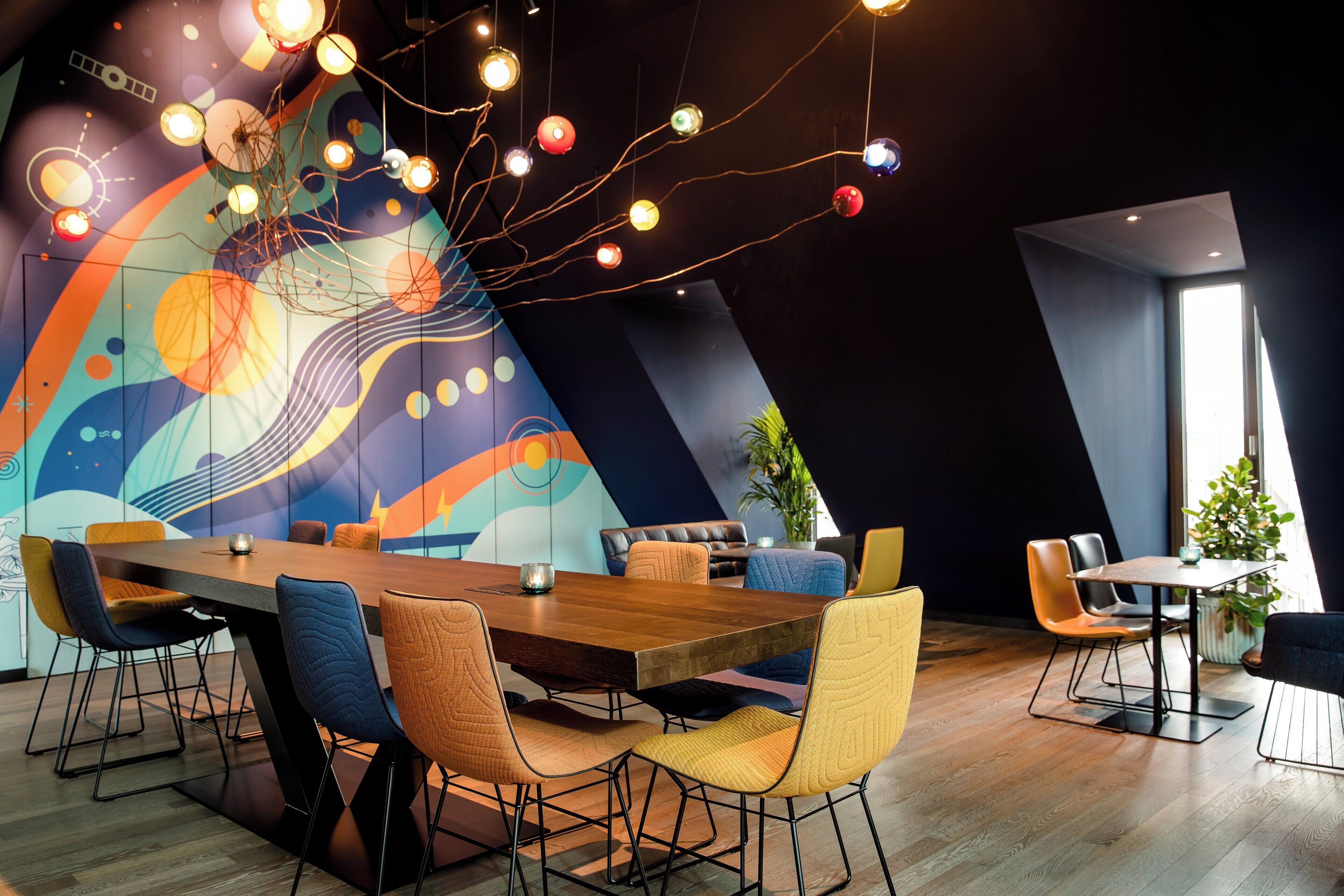 Uma zona de refeições moderna com uma mesa de madeira comprida, cadeiras coloridas e um mural abstrato vibrante na parede.