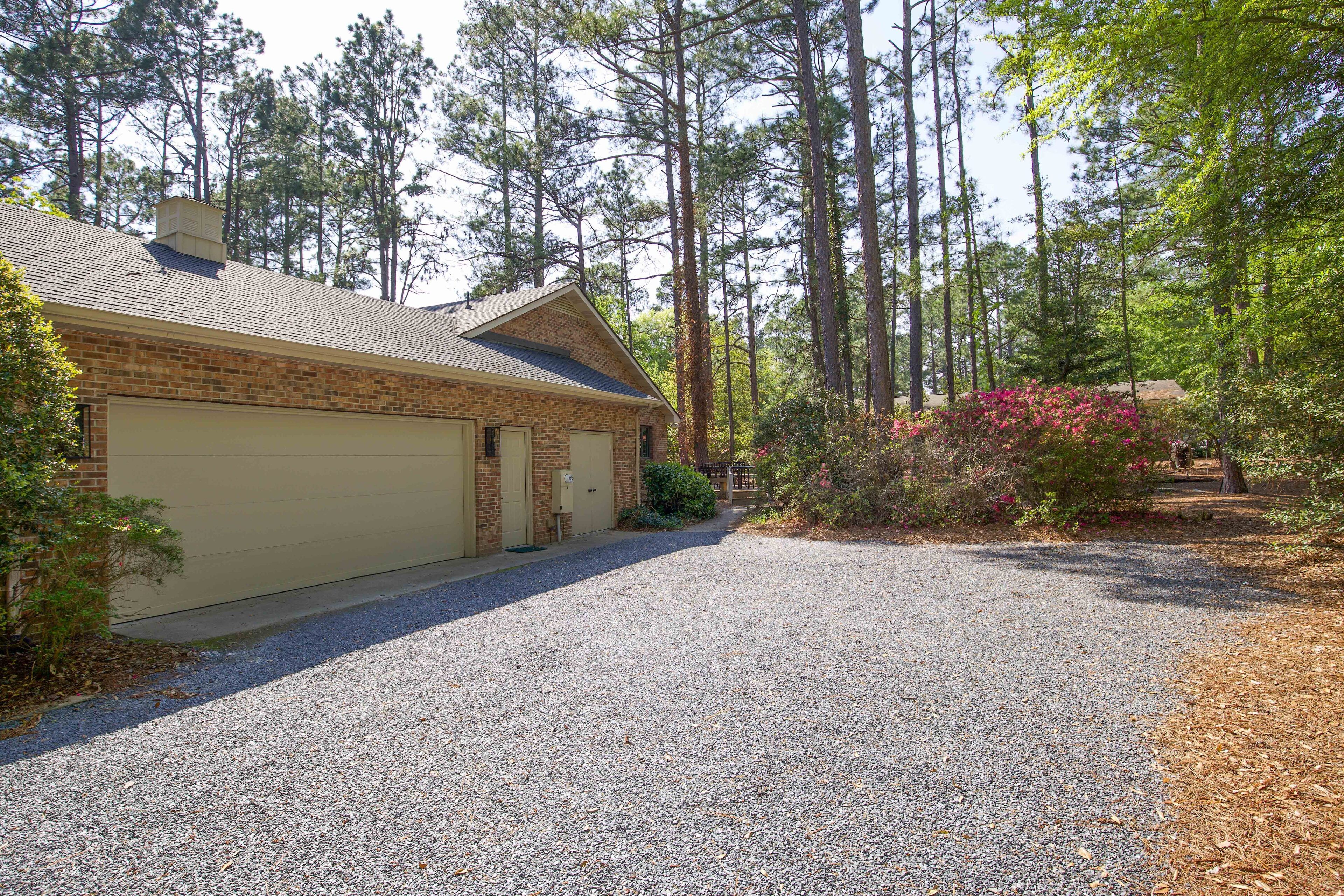 A single-story house with a gravel driveway, surrounded by tall pine trees and a small garden with pink flowers.