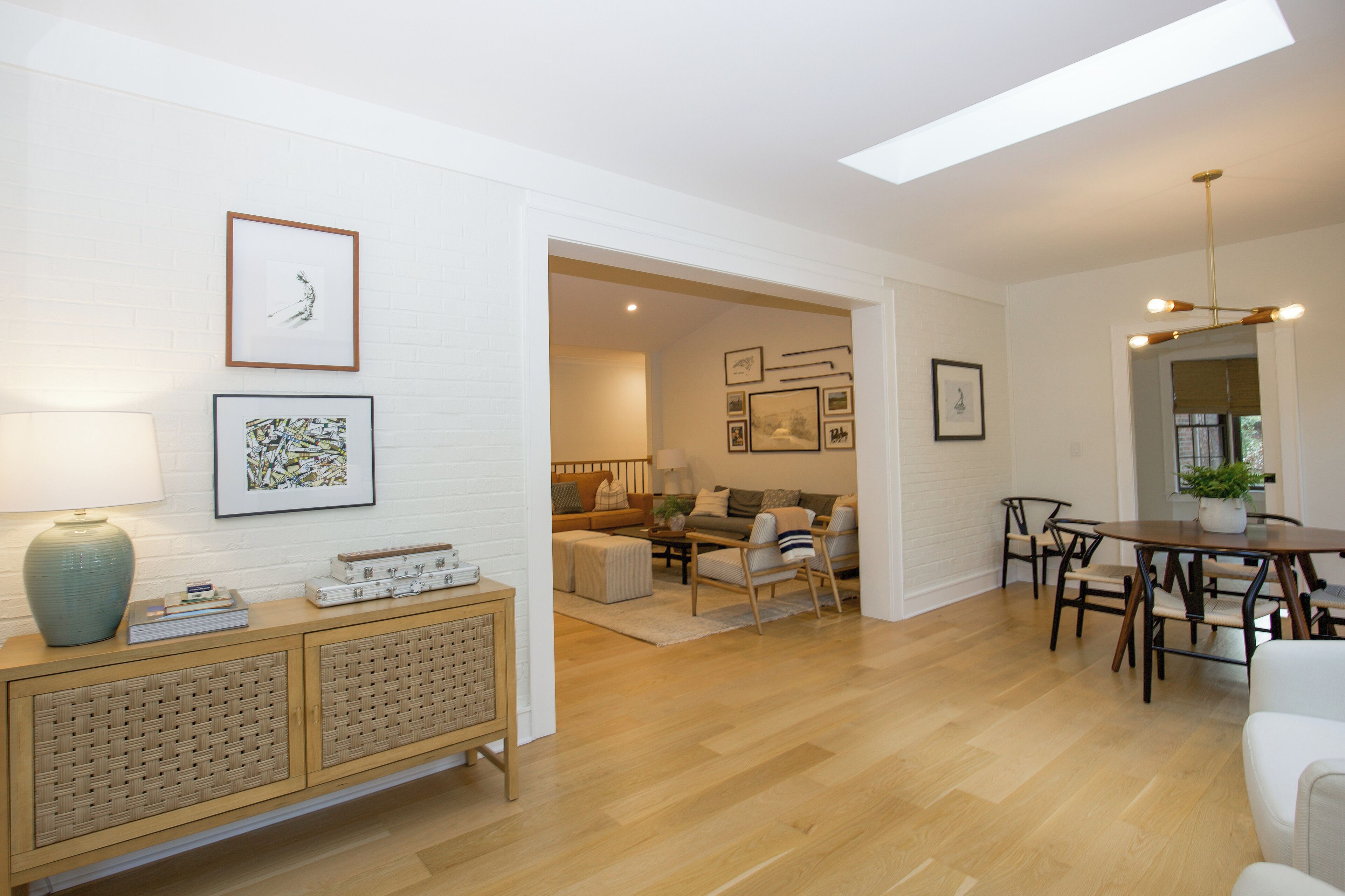 Un salon moderne avec un plancher en bois, un coin repas avec une table ronde et des chaises, et un mur en briques blanches orné de tableaux encadrés.