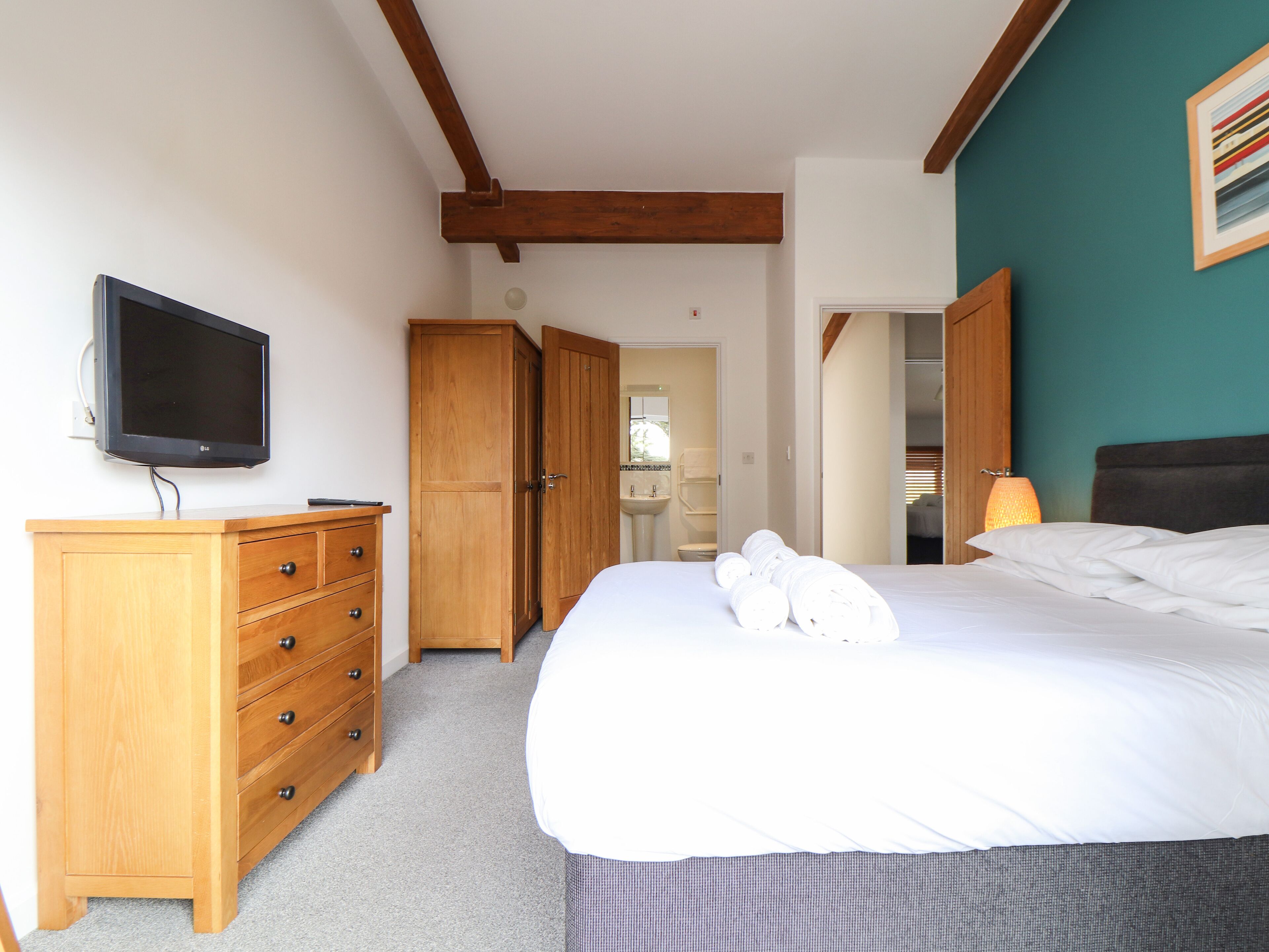 Un dormitorio con cama, cómoda de madera, televisor y baño visible a través de una puerta abierta.