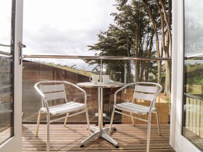 A glass-enclosed patio with two metal chairs and a small table, featuring a view of trees and a wooden fence. - 11 Towan Valley (Truro)