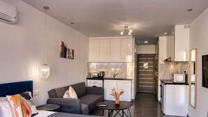 A modern living room with a sofa, a coffee table, and a TV. A kitchen with a marble backsplash and a refrigerator is visible in the background. - Phaedrus Living Seaside Luxury Flat Athina 118 (Paphos)