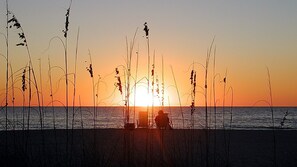 A sunset over a beach with tall grasses in the foreground and a chair facing the sunset. - Crystal Palms 201 Newly furnished,steps to beach (Treasure Island)