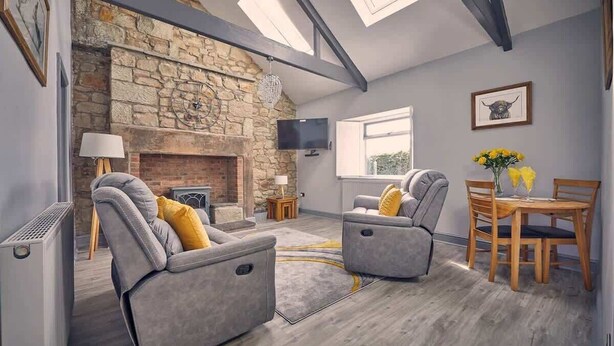 A living room with a stone fireplace, grey sofas, a dining table with chairs, and a TV mounted on the wall. - Host & Stay | Bluebell Cottage (Belford)