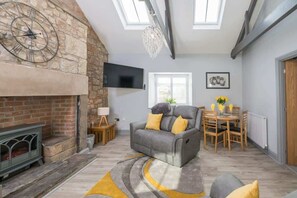 A living room with a stone fireplace, a grey sofa with yellow cushions, a dining table with chairs, and a flat-screen TV mounted on the wall.