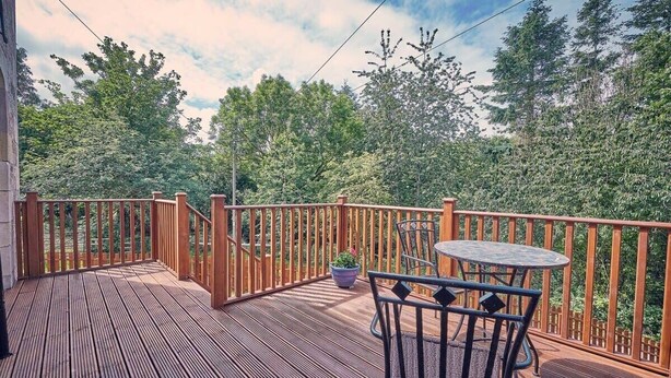 A wooden deck with a round table and chairs, surrounded by a wooden railing, overlooking a forested area.