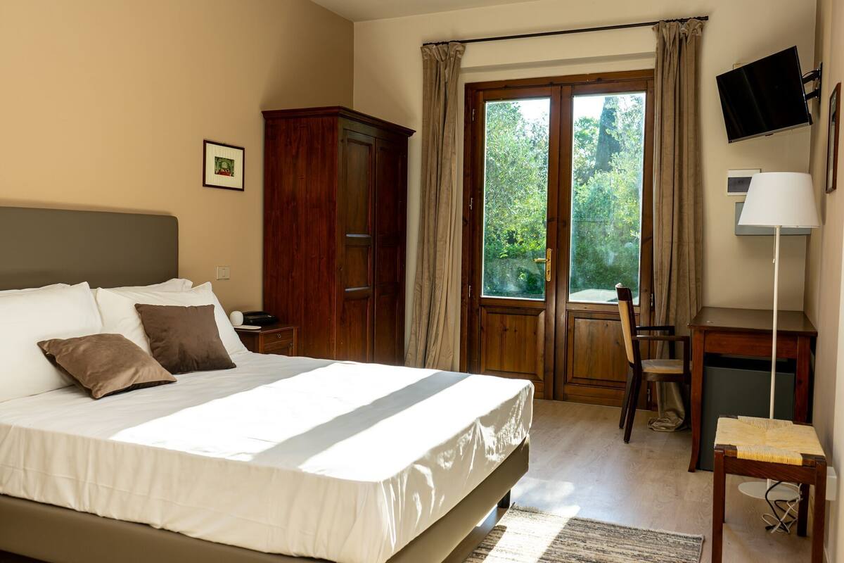 A bedroom with a bed, a desk, a chair, a TV, and a window with curtains.