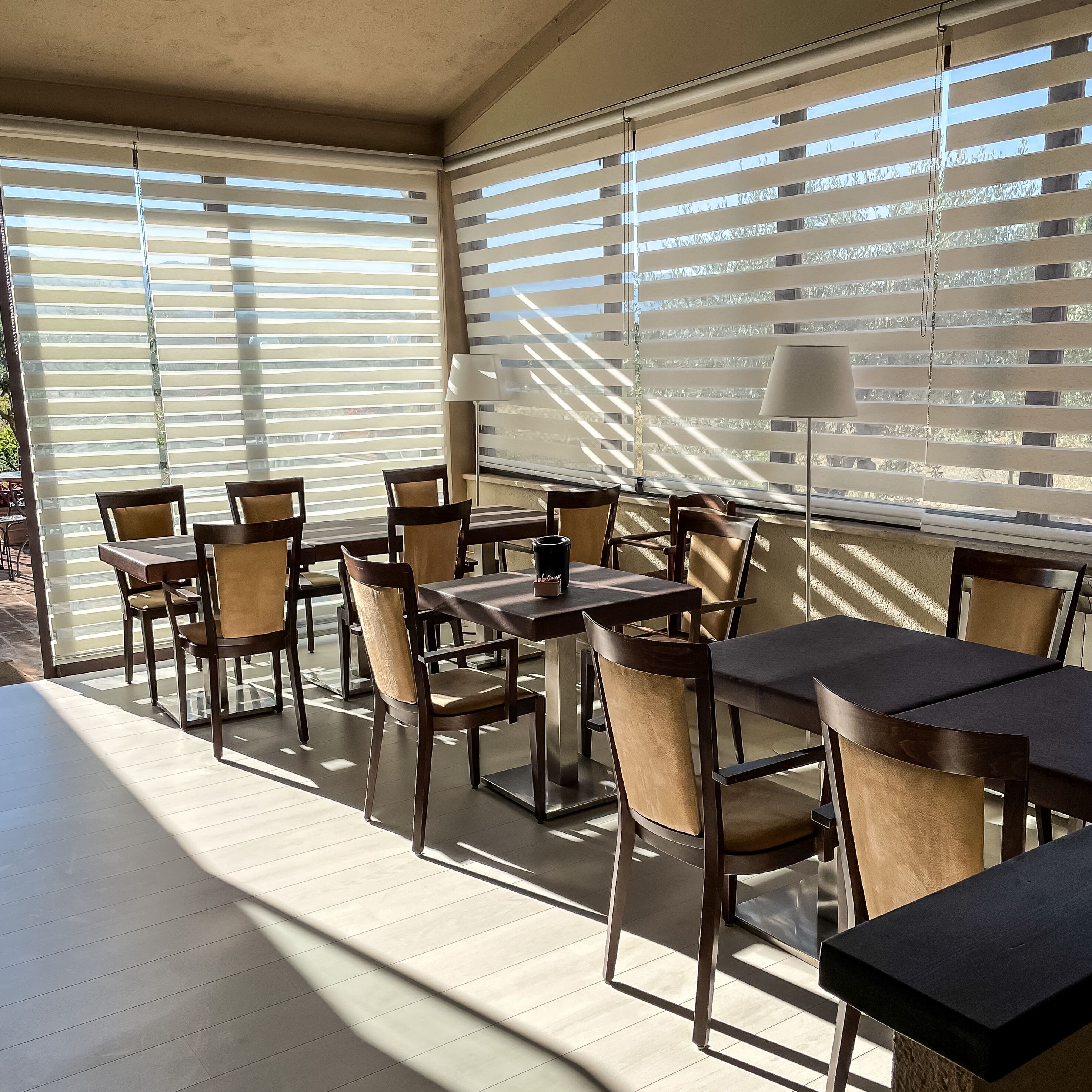A sunlit patio area with tables and chairs, large windows with blinds, and a lamp.