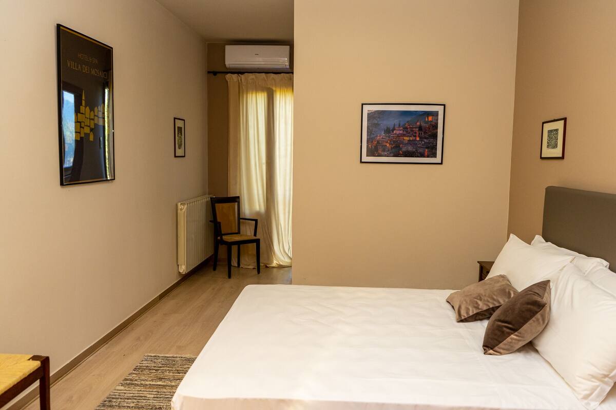 A hotel room with a large bed, a chair, a radiator, and a window with curtains.