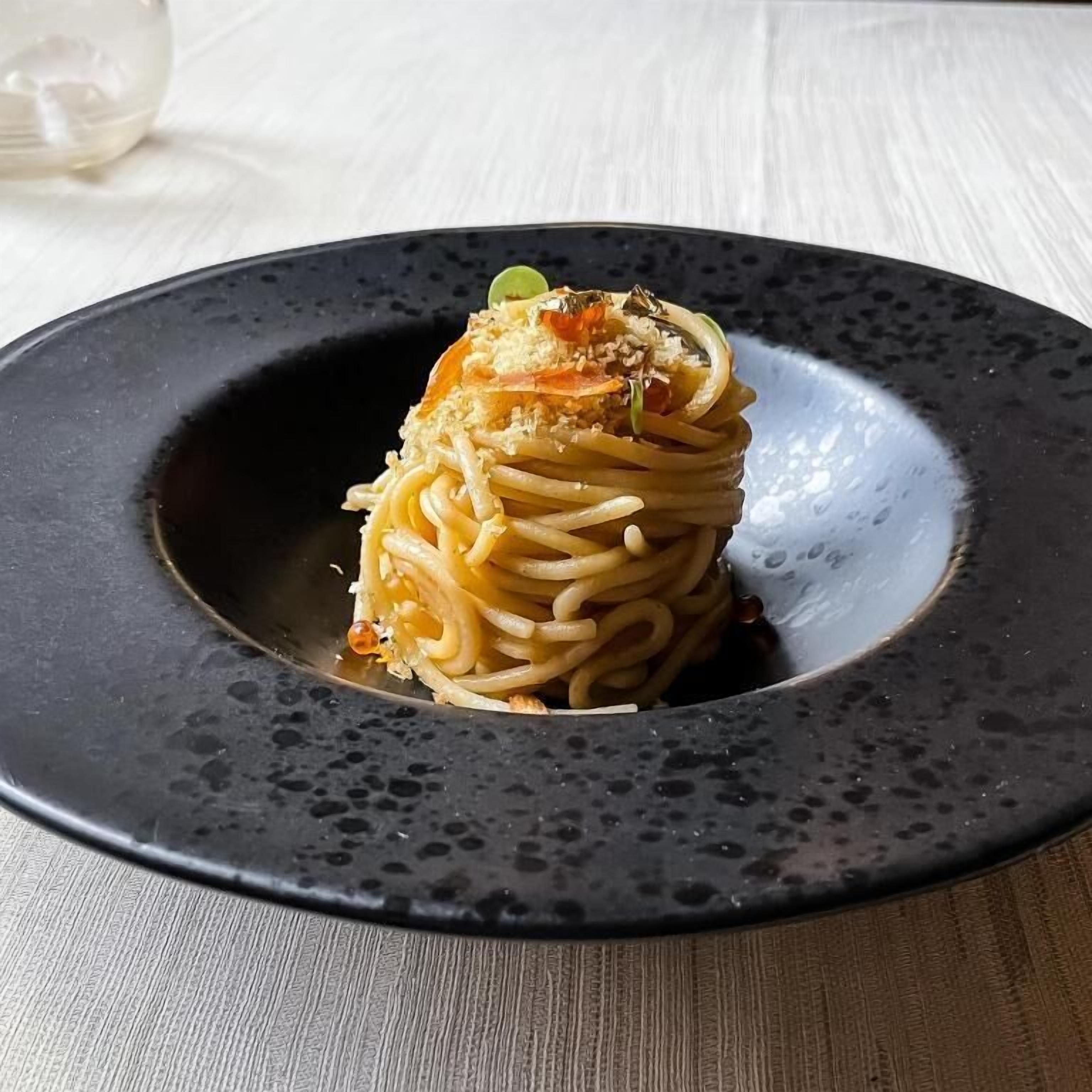 Un piatto di spaghetti con formaggio e verdure, servito su un piatto scuro e maculato.