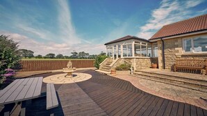 A wooden deck with a circular seating area, a stone staircase, and a covered outdoor area with a view of the sky. - Host & Stay | Cheviot View (Seahouses)