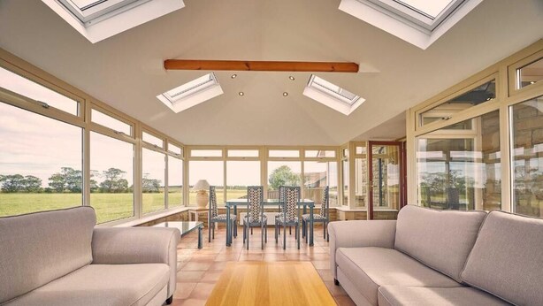 A sunroom with a wooden floor, two sofas, a dining table with chairs, and large windows. - Host & Stay | Cheviot View (Seahouses)