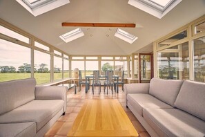 A sunroom with a wooden floor, two sofas, a dining table with chairs, and large windows.