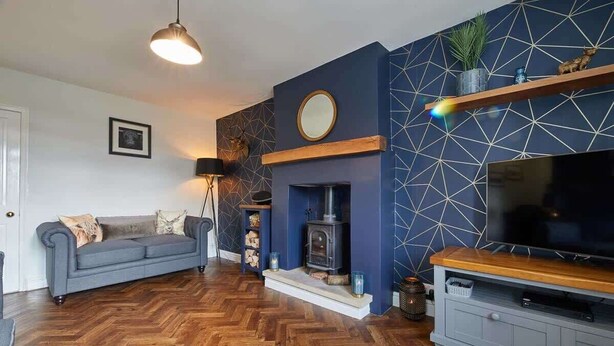 Una sala de estar con piso de madera en patrón chevron, un sofá gris, una chimenea, un televisor sobre un mueble de madera y una pared con diseño geométrico.