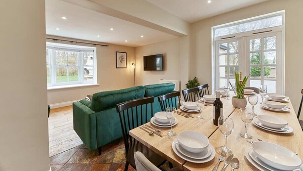 A dining area with a wooden table set for four, a green sofa, and a television mounted on the wall. - Host & Stay | The Old Rectory (Witton Gilbert)