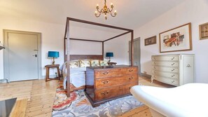 Een hemelbed met houten frame, een dressoir met meerdere laden, een kroonluchter en een abstract schilderij in een lijst aan de muur.
