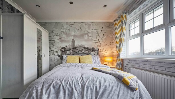 Un dormitorio con un mural de paisaje urbano en la pared, una cama con ropa de cama estampada, una mesita de noche con una lámpara y una ventana con cortinas.