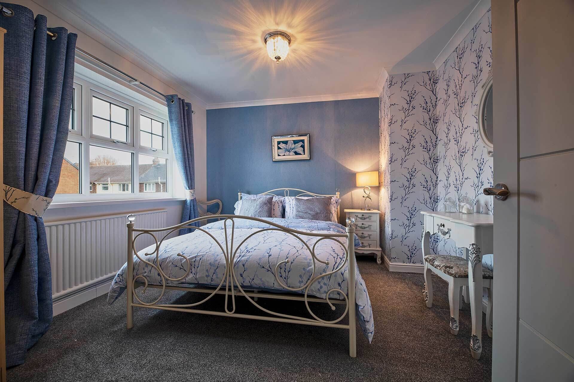 Un dormitorio con cama, mesita de noche, lámpara, ventana con cortinas y un papel tapiz con estampado floral.