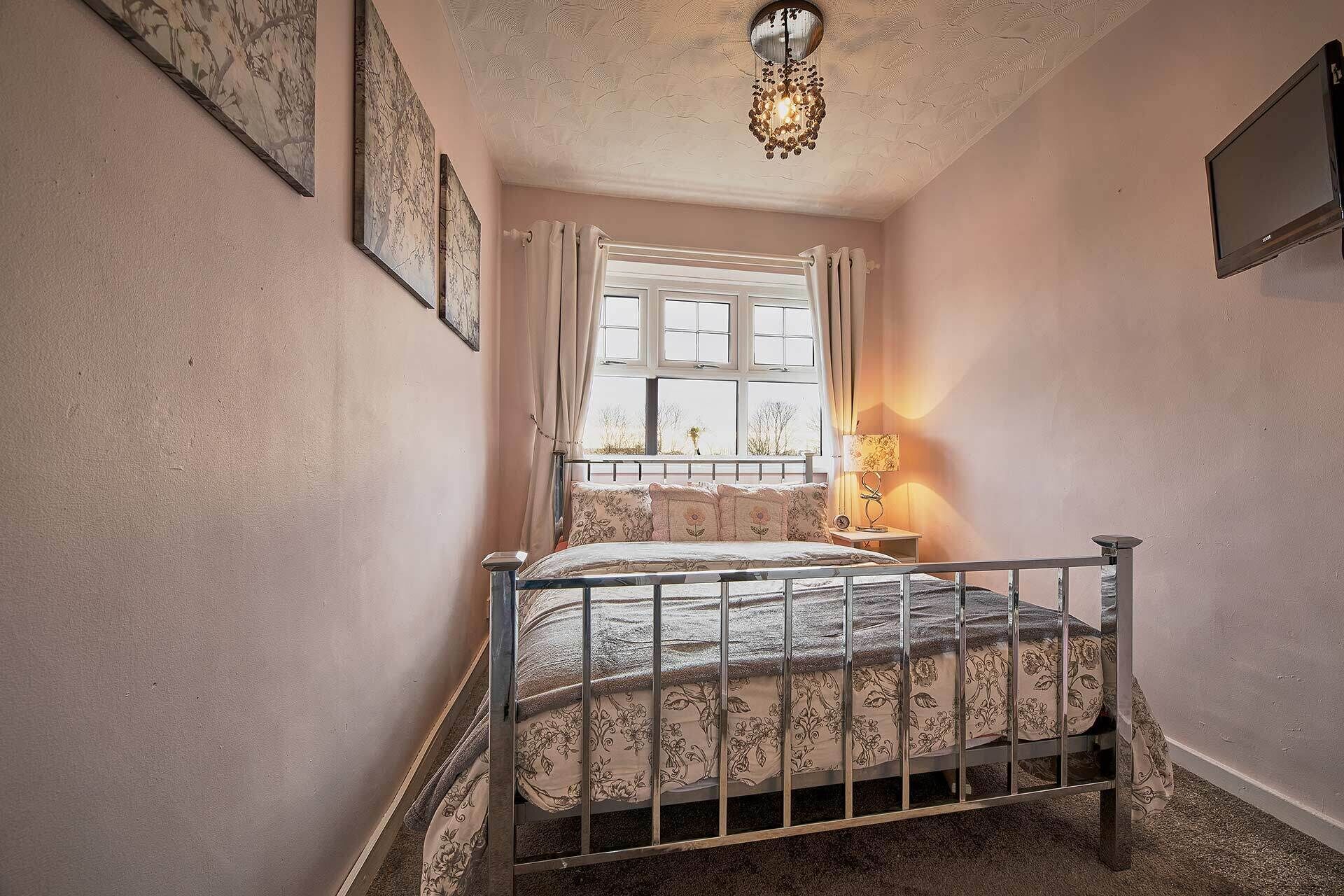 Une chambre à coucher avec un lit en métal, une fenêtre avec des rideaux, un téléviseur fixé au mur et un luminaire décoratif au plafond.