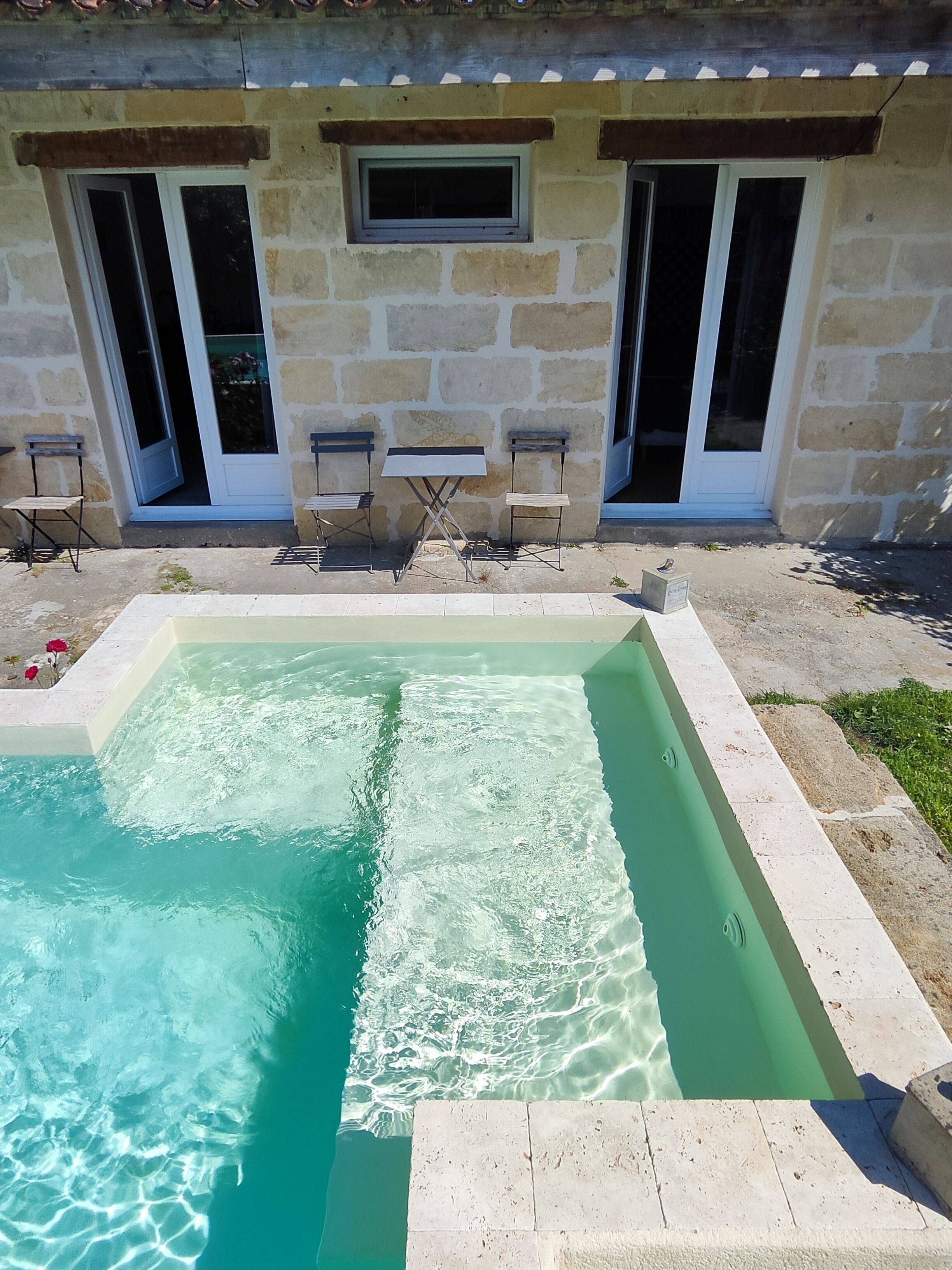 A swimming pool with a stone border, two chairs, and a stone wall with a window.