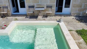 A swimming pool with a stone border, two chairs, and a stone wall with a window.