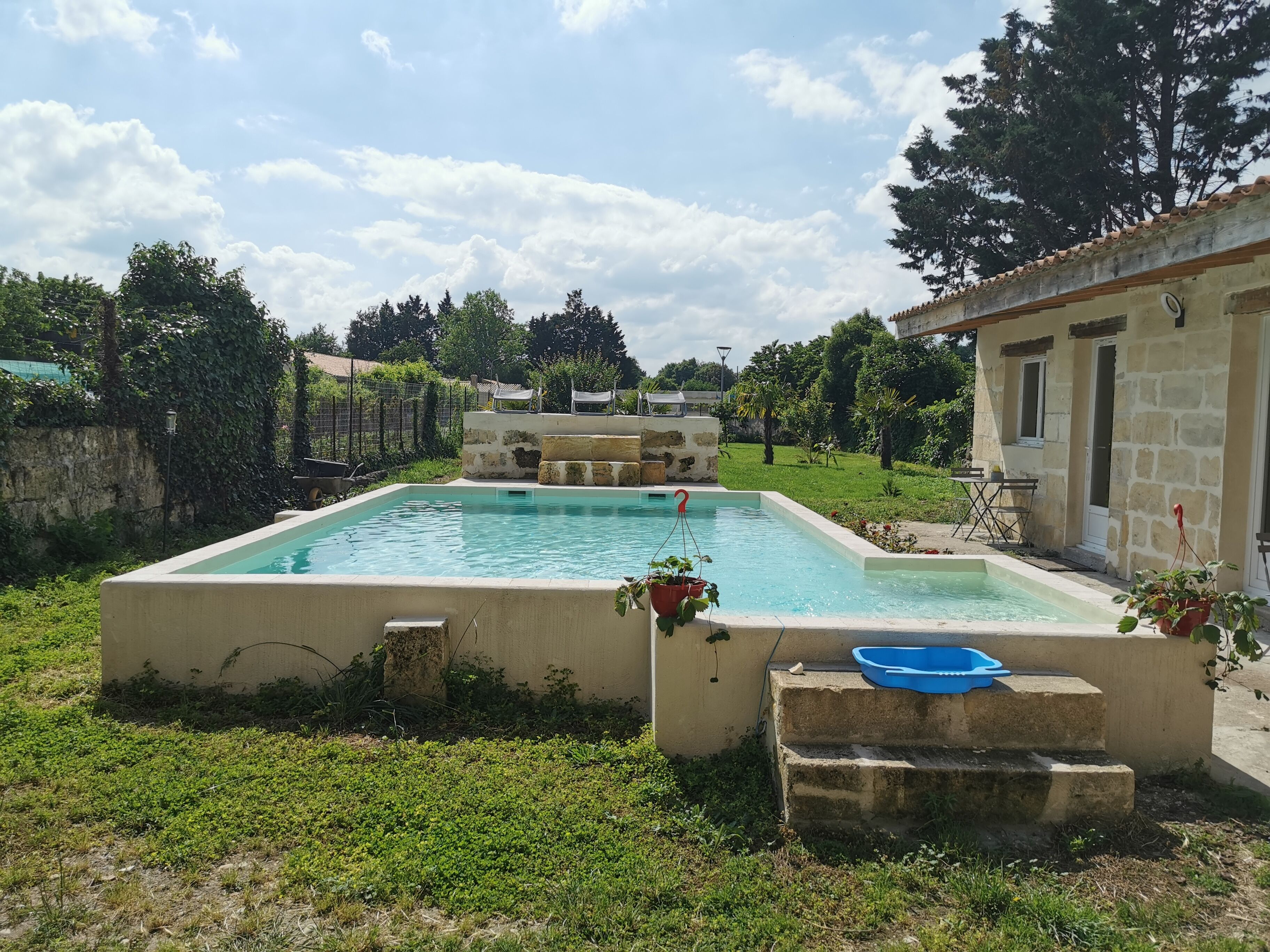 Ein rechteckiger Pool mit Stufen, die zu einer kleinen Terrasse führen, umgeben von viel Grün und einem Steinhaus.