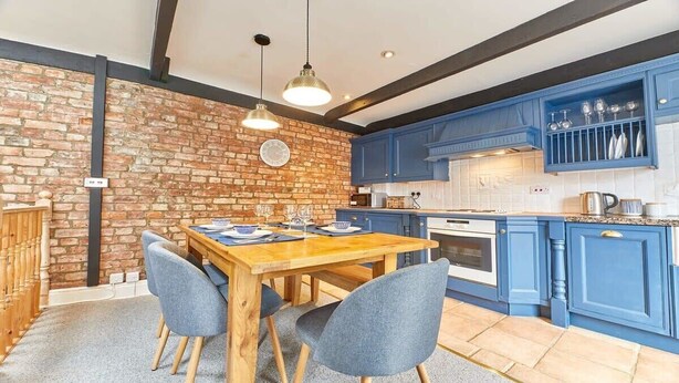 A kitchen with blue cabinets, a brick wall, a wooden dining table, and chairs. - Host & Stay | The Burrow (Alnwick)