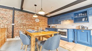 A kitchen with blue cabinets, a brick wall, and a wooden dining table with chairs. - Host & Stay | The Burrow (Alnwick)