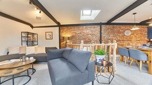 A modern living room with a grey sofa, a wooden coffee table, and a dining area with a brick wall and wooden furniture. - Host & Stay | The Burrow (Alnwick)