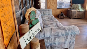 A cozy room with a 'Welcome' sign, a rustic wooden door, a patterned sofa, and a wooden cabinet with a television. - Sweet Riverside Cabin, Easy Walk 2 River, Private River Access, River Views! (Concan)
