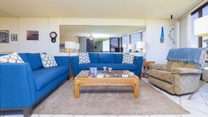 Una sala de estar con un sofá azul, un sofá color beige, una mesa de centro de vidrio y un espejo grande que refleja la habitación.