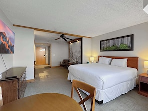A hotel room with a bed, a desk, a chair, a ceiling fan, and a painting on the wall.