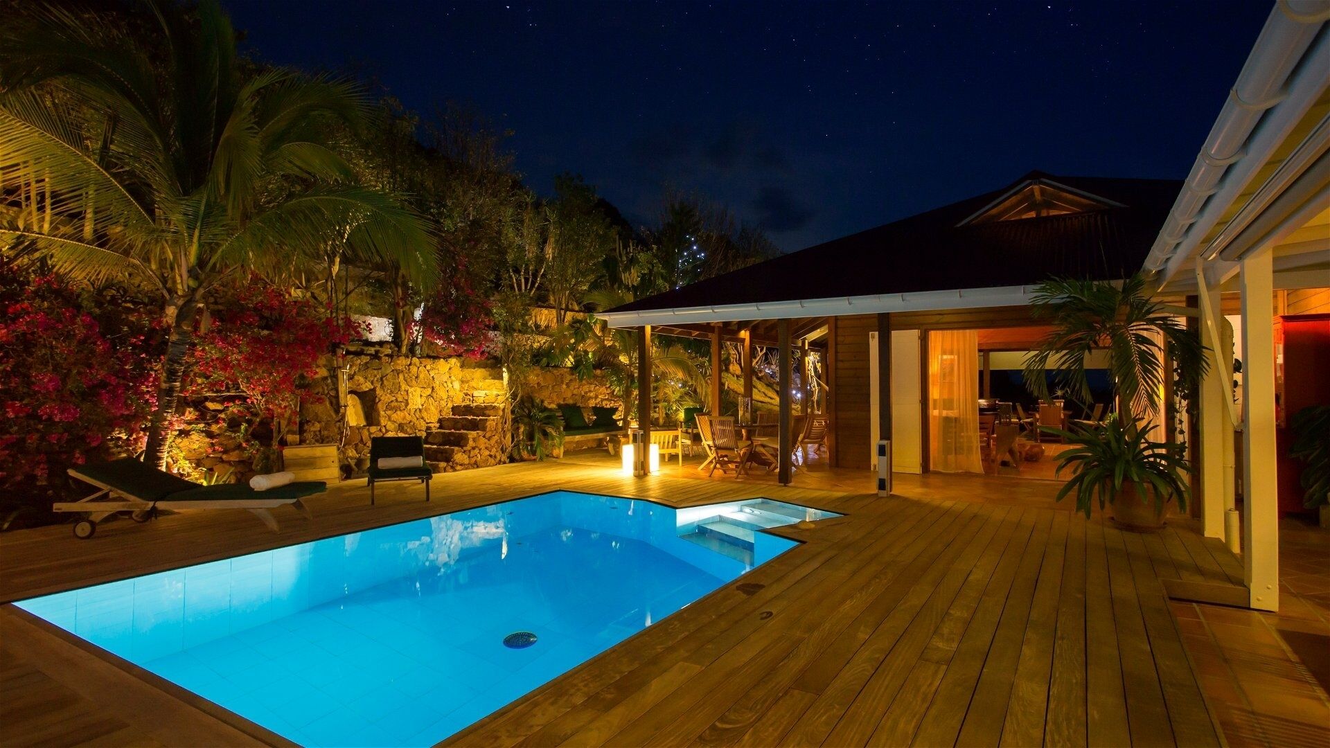 A nighttime view of a pool area with a wooden deck, lounge chairs, and a stone wall.