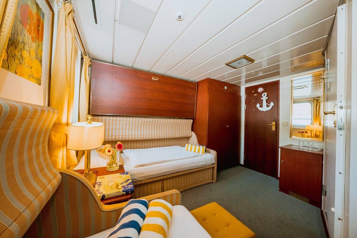 A cozy cabin with a bed, a small table with a lamp, a window, and a door with an anchor decoration.