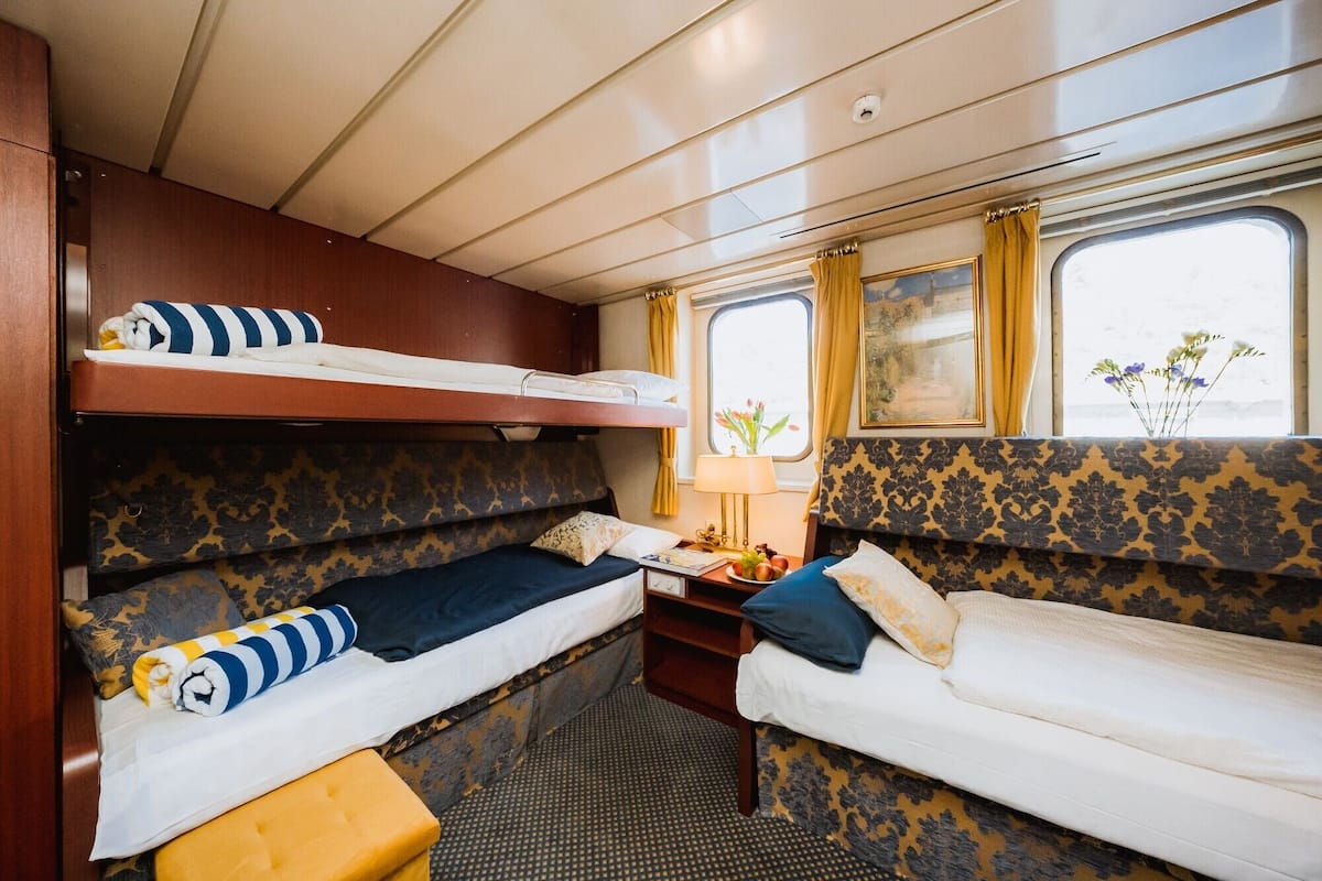 A compact train cabin with two bunks, a small table with a lamp, and a window with curtains.