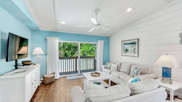 A modern living room with a white sofa, a coffee table, and a TV on a white cabinet. There is a balcony with white railings and a view of greenery. - Anna Maria Home with Private Pool, Near the Beach! - Coveside Cottage (Holmes Beach)