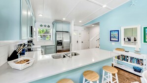 A modern kitchen with white countertops, a stainless steel refrigerator, and a sink. There are three stools at the counter and a bookshelf with books. - Anna Maria Home with Private Pool, Near the Beach! - Coveside Cottage (Holmes Beach)