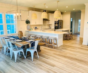 A modern kitchen with a central island, pendant lights, and a dining area with white chairs.