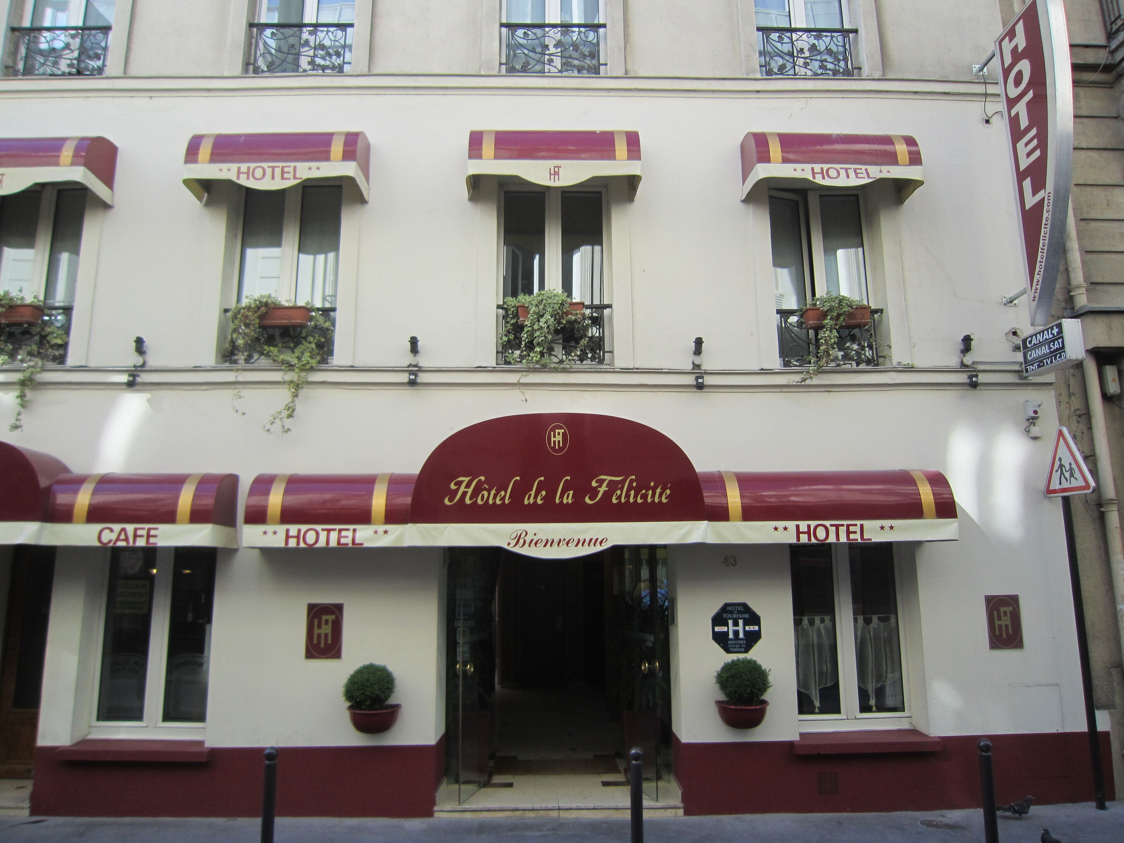 A building with multiple "HOTEL" signs and a "CAFE" sign. There are potted plants on the balconies and a pedestrian crossing sign.
