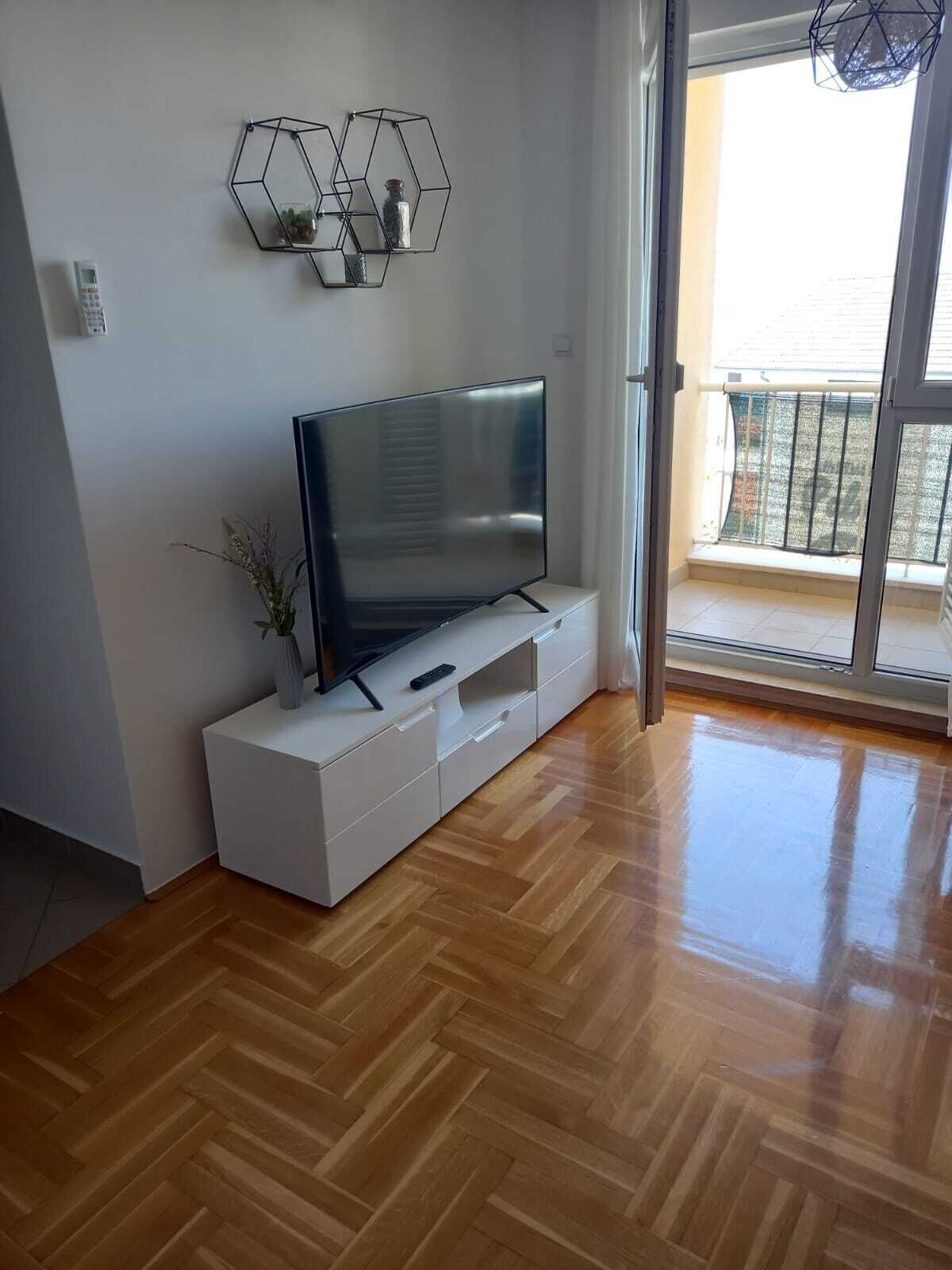 Una sala de estar moderna con un televisor de pantalla plana sobre un mueble blanco, suelo de parquet de madera y un balcón con vistas.