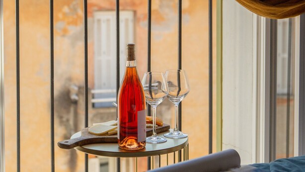 Ein Balkon mit Tisch, darauf eine Flasche und Gläser, ein Fenster mit Vorhängen und Blick auf ein Gebäude mit roten Ziegeln.