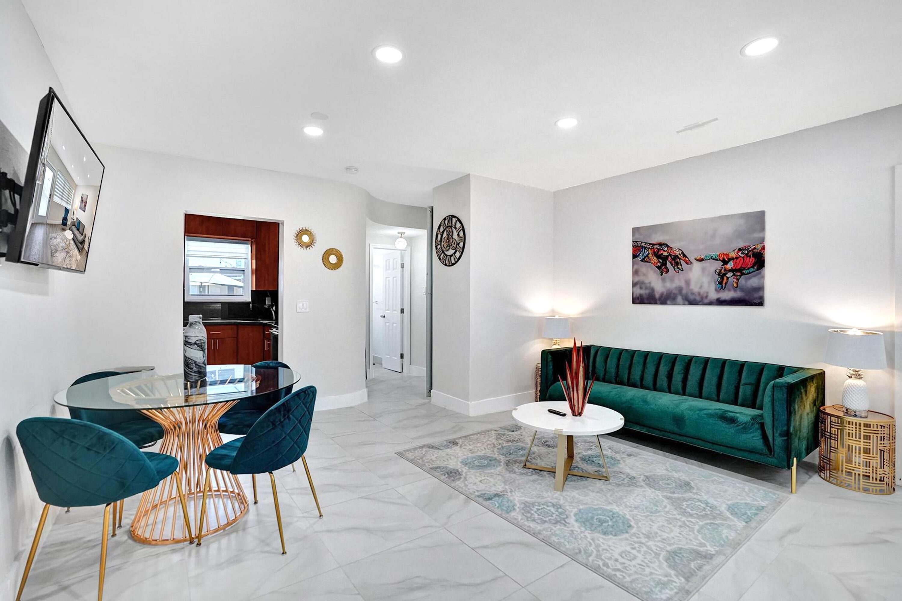 A modern living room with a glass dining table, teal chairs, a green sofa, and a patterned rug.