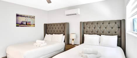 A hotel room with two beds, a ceiling fan, an air conditioning unit, and a painting on the wall.