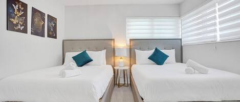 A hotel room with two beds, white bedding, teal pillows, a small bedside table with a lamp, and artwork on the wall.